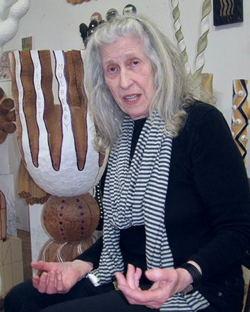 Sculptor Linda Brenner speaks at a lecture, gesticulating with her hands while she sits in front of her sculptures