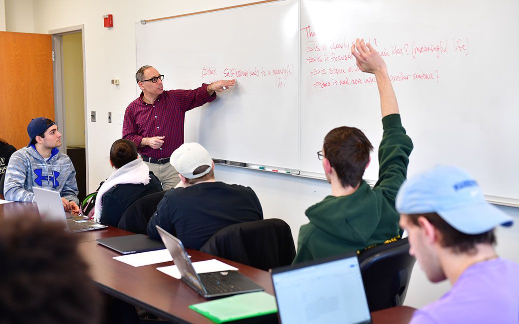 Class meeting in a classroom, with a professor speaking to students seated and raising hands