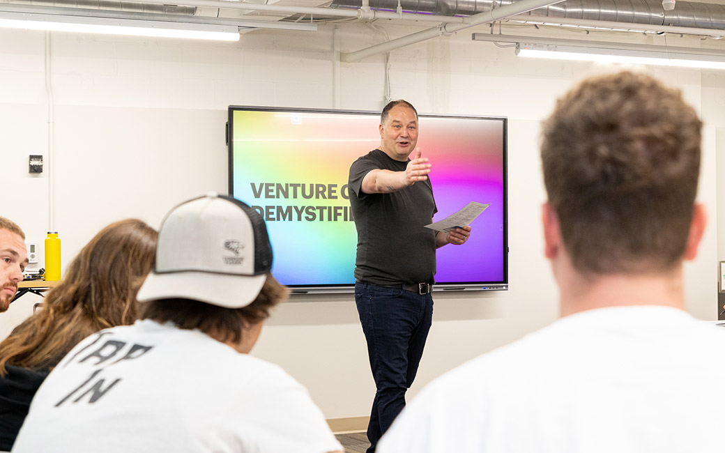 Man presents in conference room with screen "Venture Creation Demystified"