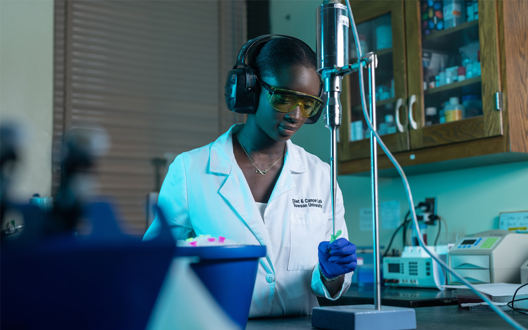 Zeguela Kamagate stands in a white lab coat and wearing personal protective equipment for her eyes and ears. She is pictured holding a test tube under a tool called a sonicator.